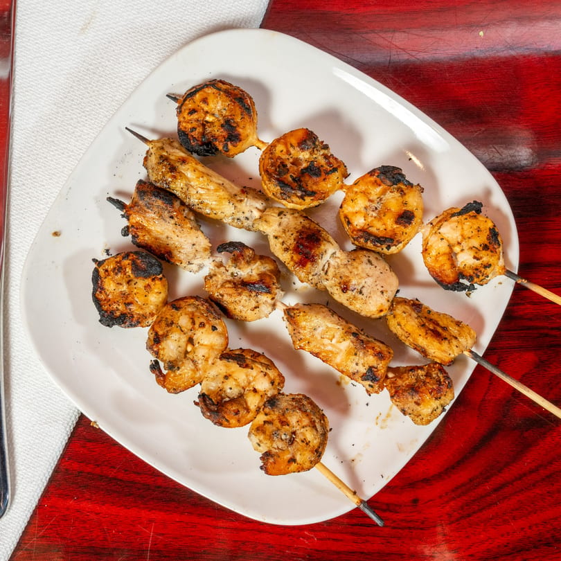 Grilled chicken and shrimp skewers on a white plate, with a red wooden table background.