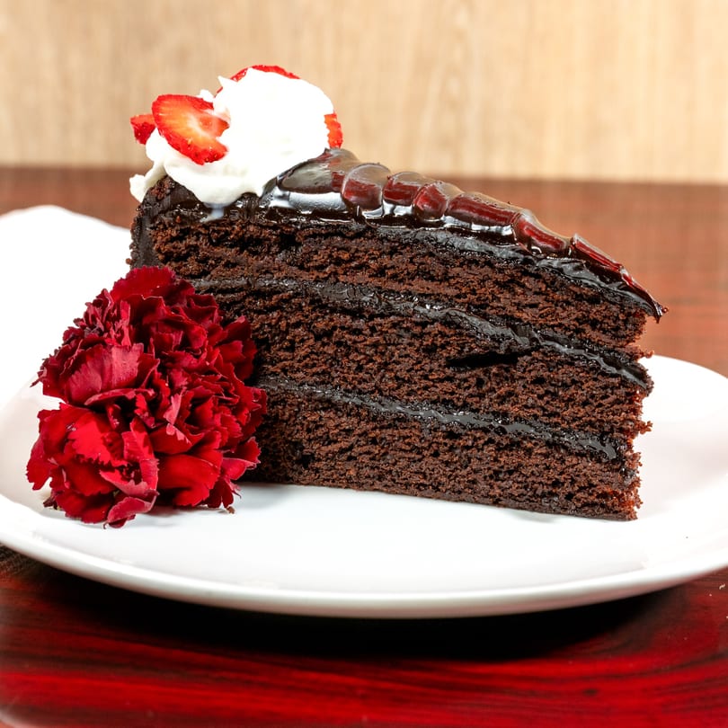 Slice of chocolate cake with whipped cream, strawberries, and red flowers on a white plate.