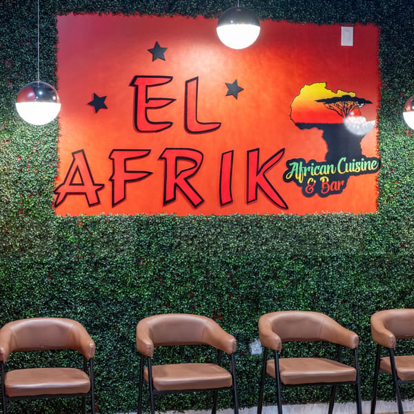 El Afrik Cuisine and Bar sign on a red background with a green leafy wall and brown chairs.