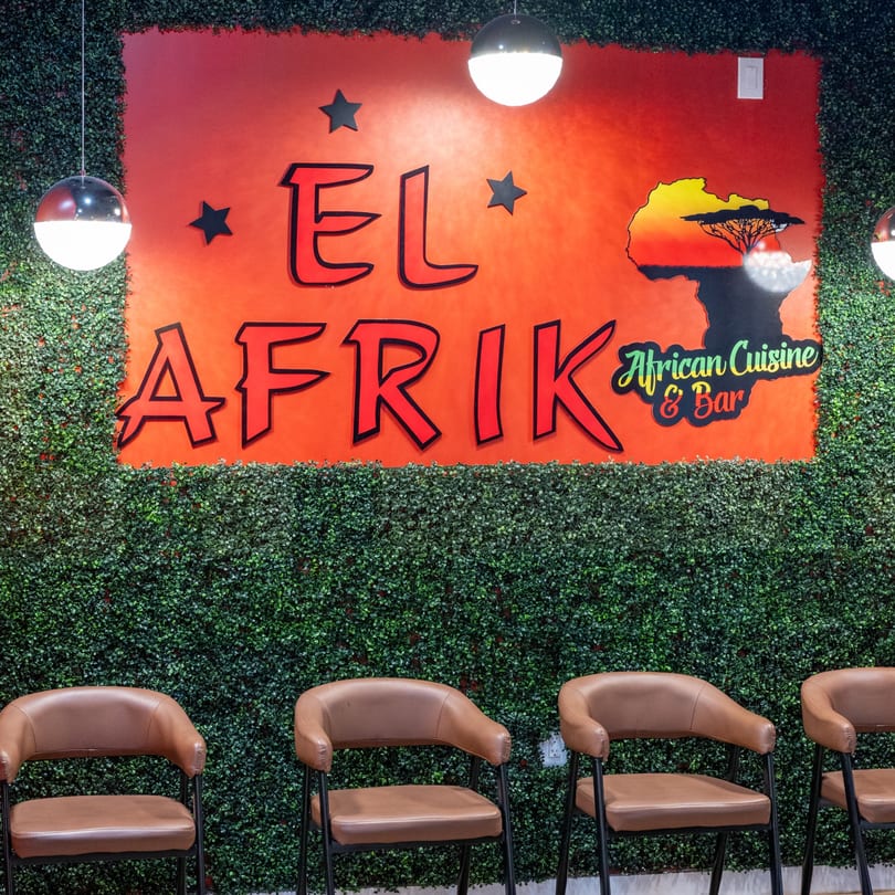 El Afrik Cuisine and Bar sign on a red background with a green leafy wall and brown chairs.