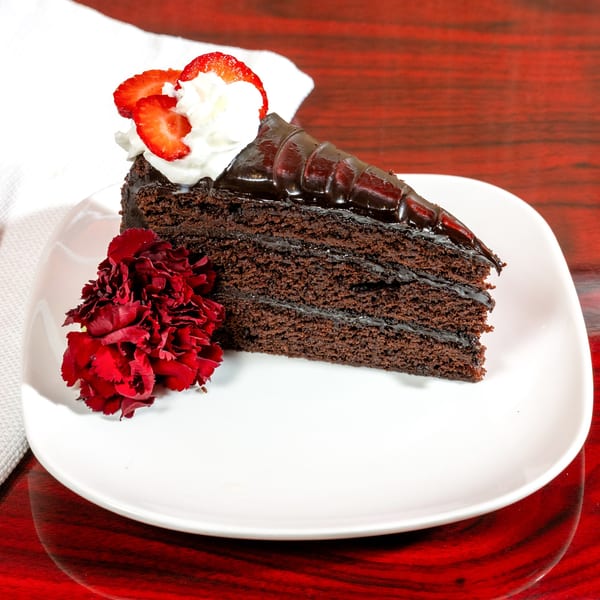 Slice of chocolate cake with whipped cream, strawberries, and a red flower on a white plate.