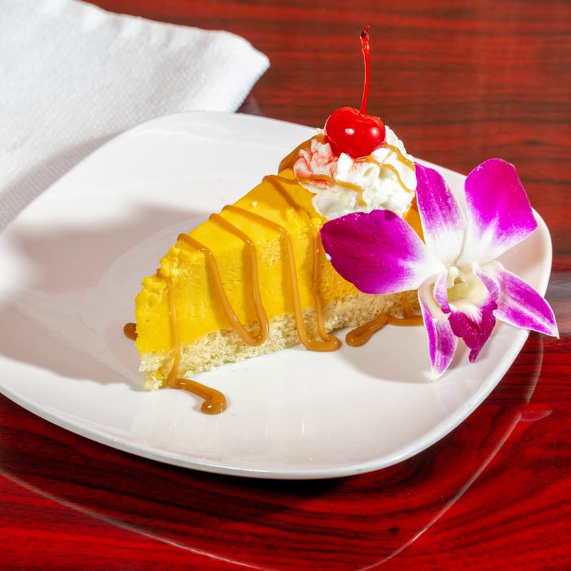 Slice of mango cheesecake with caramel drizzle, whipped cream, a cherry, and an orchid.