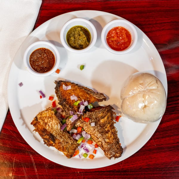Fried fish with chopped vegetables, three dipping sauces, and fufu on a white plate.