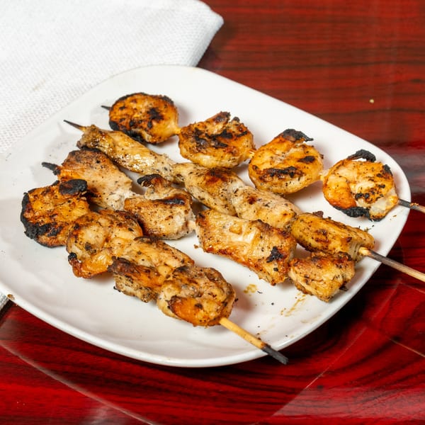 Grilled chicken and shrimp skewers with char marks on a white plate, on a red wooden table.