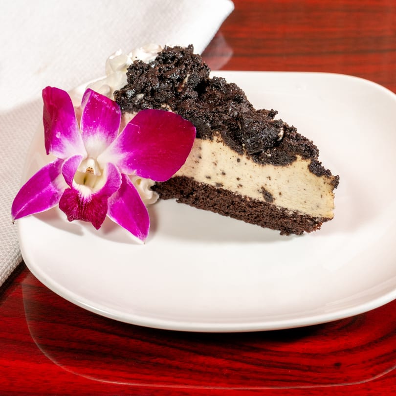 A slice of cookies and cream cheesecake with whipped cream and a purple orchid on a white plate.