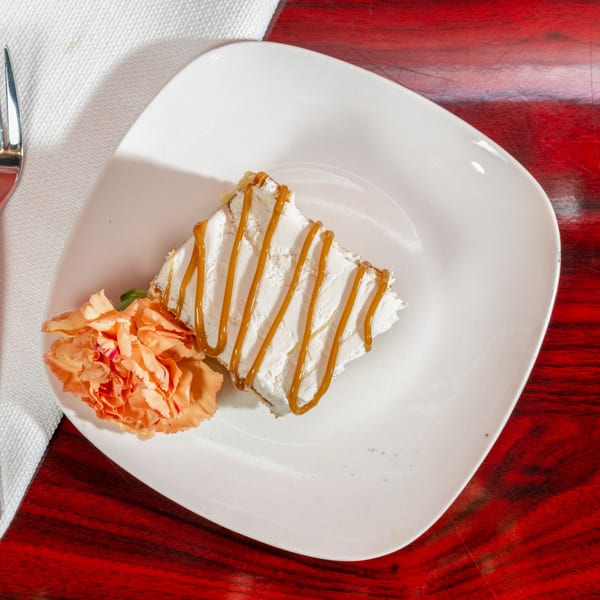 A slice of white cake with caramel drizzle and an orange flower on a white plate.