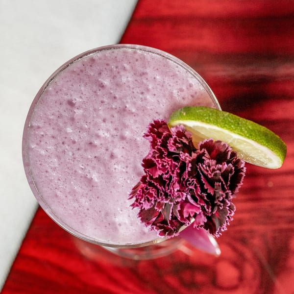 Top-down view of a foamy pink cocktail garnished with a lime wedge and purple flowers.