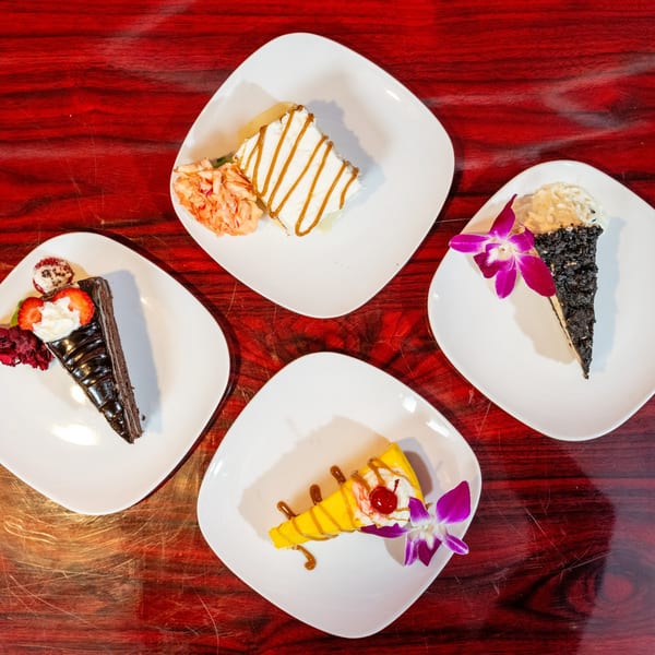 Four slices of cake, including chocolate, mango, and caramel, on white plates with floral garnishes.