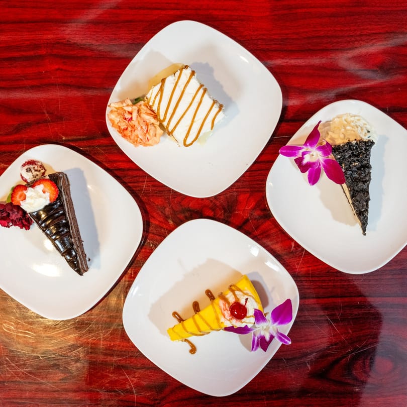 Four slices of cake, including chocolate, mango, and caramel, on white plates with floral garnishes.