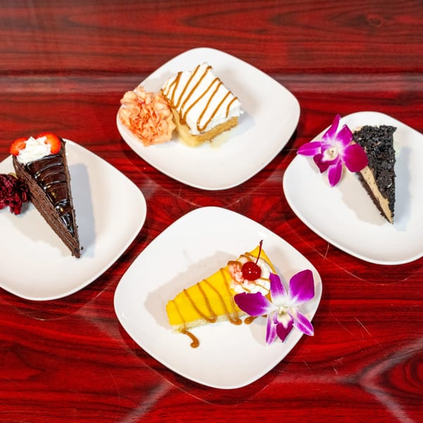 Four slices of cake, including chocolate, mango, and caramel, on white plates with floral garnishes.