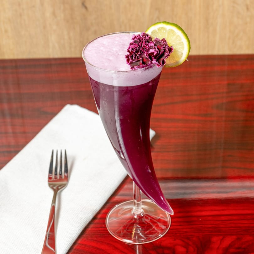 A purple drink in a unique horn-shaped glass, garnished with a lime slice and purple flower.